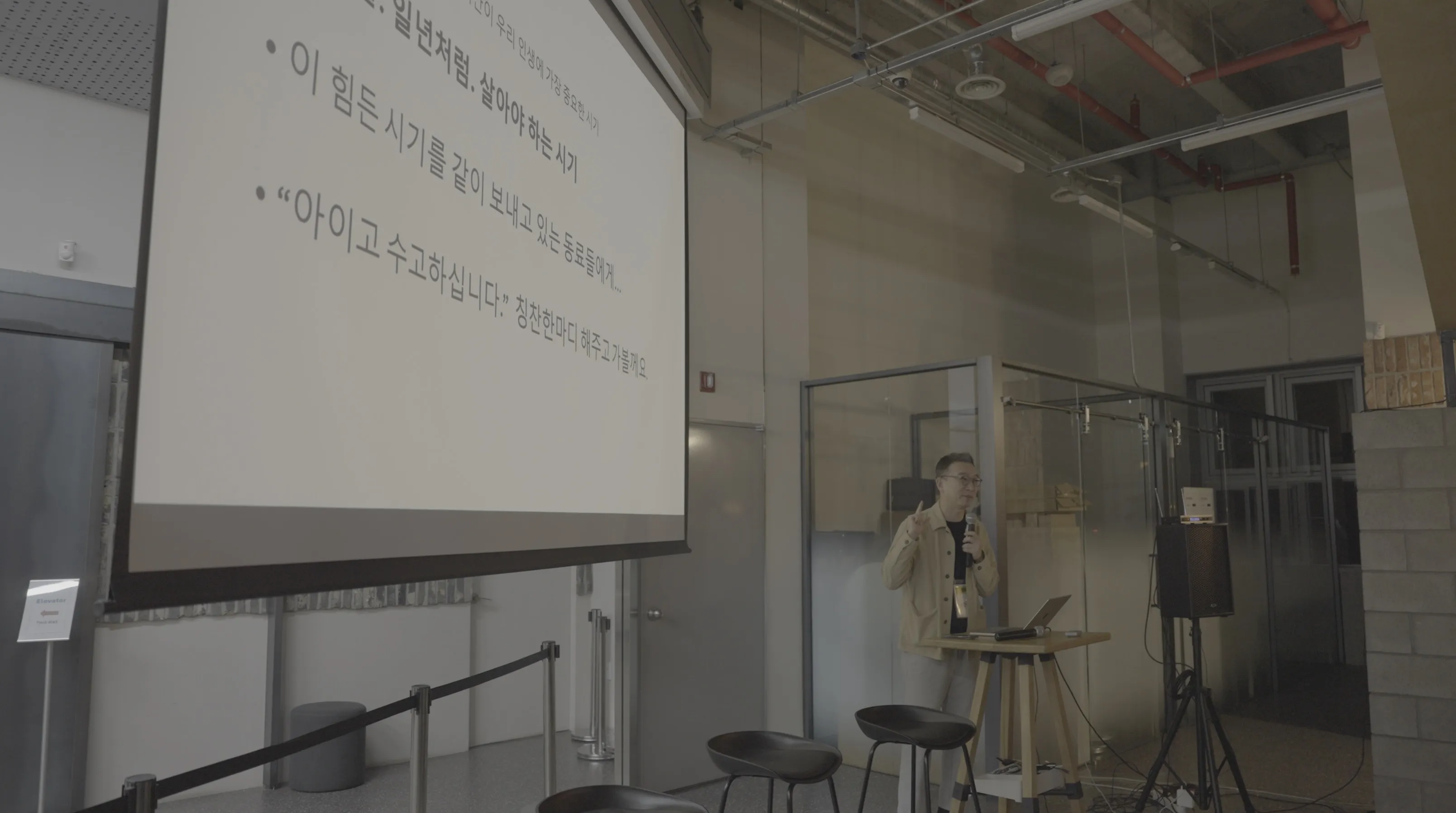 발표자가 강연 스탠드 앞에 서 있고 대형 스크린에 '이 힘든 시기를 같이 보내고 있는 동료들에게', '아이고 수고하십니다' 등의 문구가 표시된 도망자 연합 행사 오프닝 발표 장면